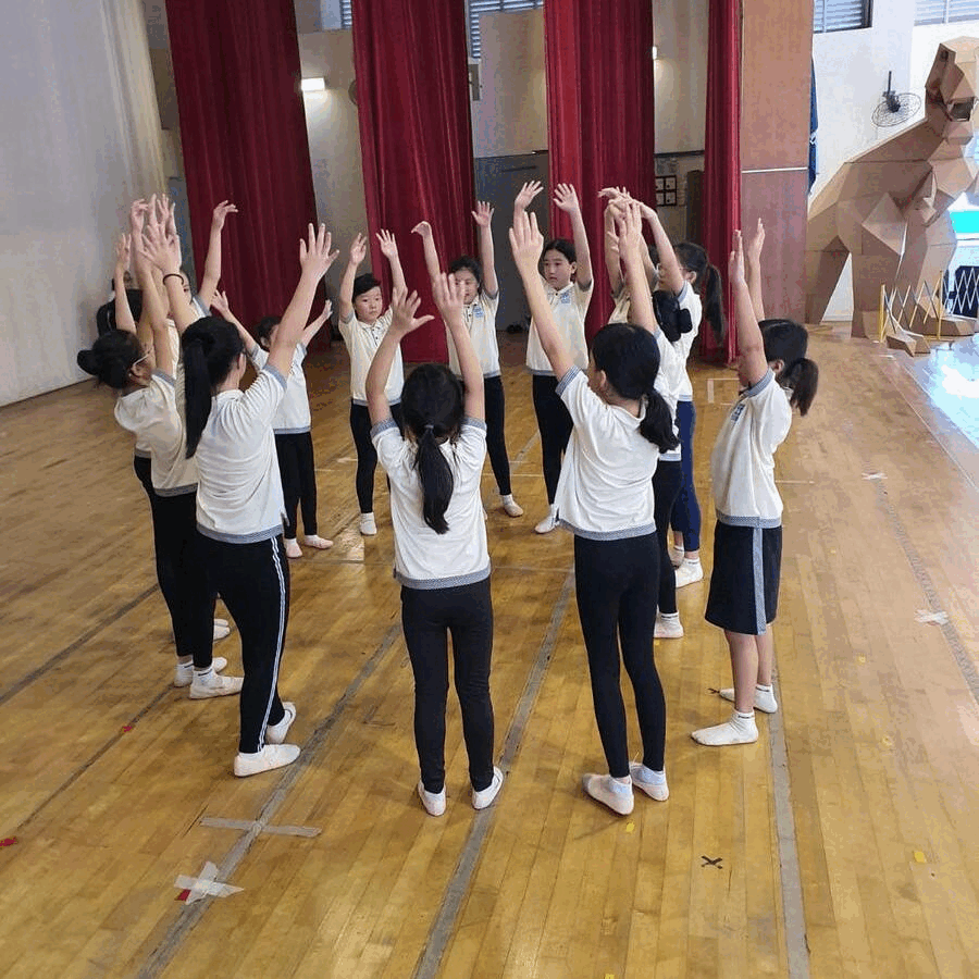 Children in white shirts raise their arms in a circle on a wooden gym floor near red curtains.